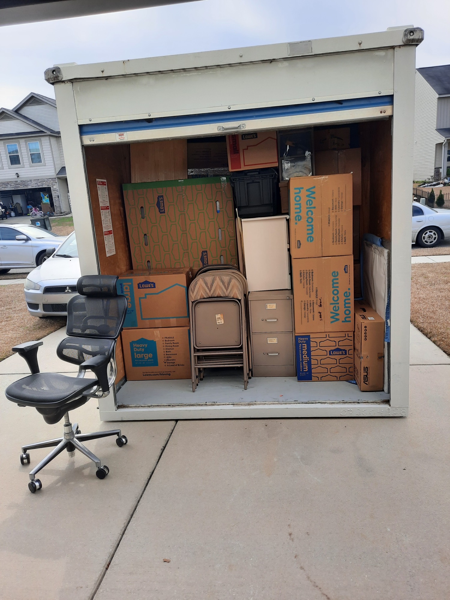 Portable moving container in a driveway loaded with labeled boxes, filing cabinets and chairs