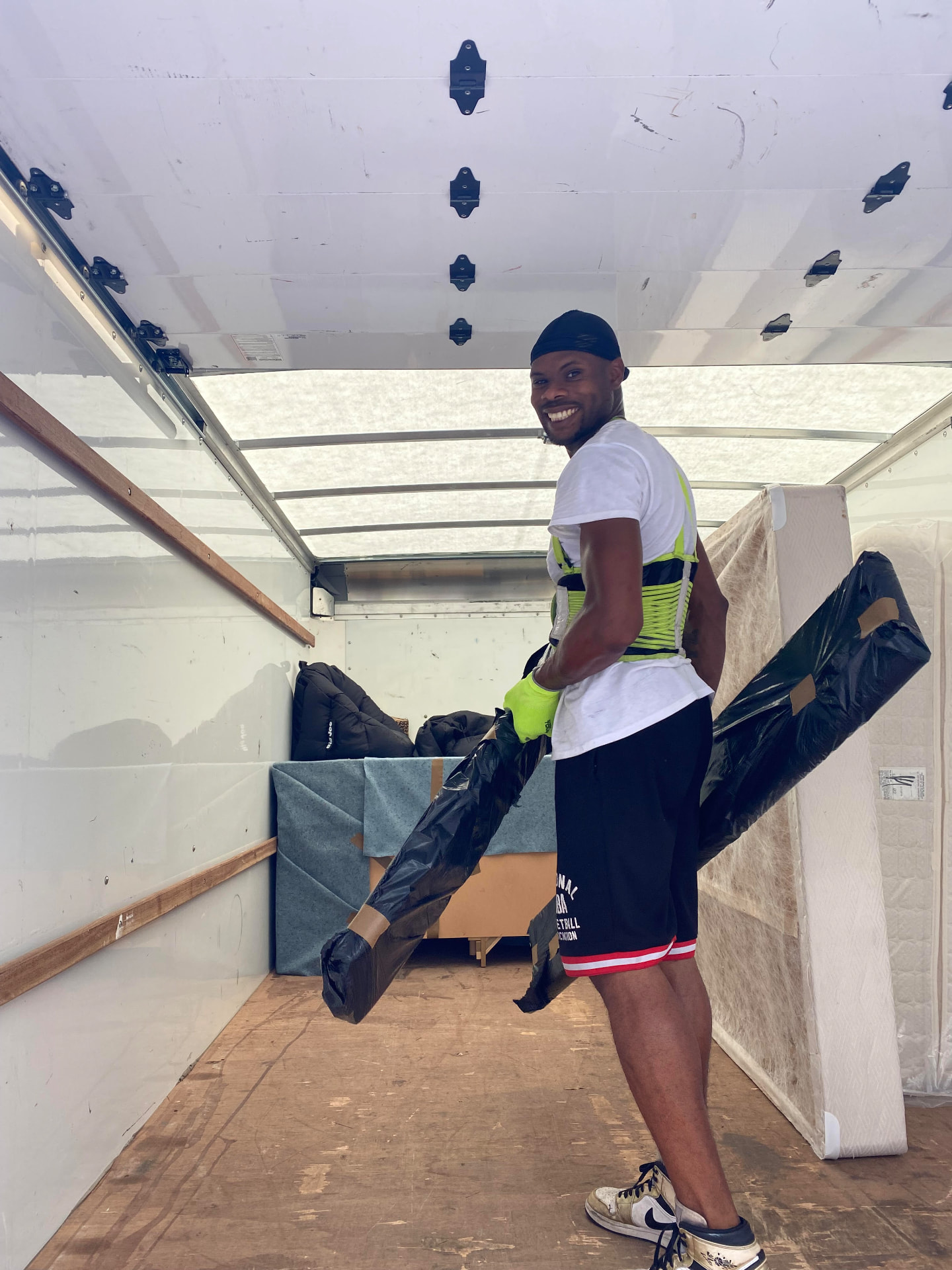 Mover in a support belt and work gloves carrying a wrapped mattress into a moving truck